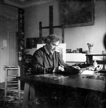 Marc Chagall in a suit, writing on the studio table, in front of the work The Cemetery hanging on the wall.