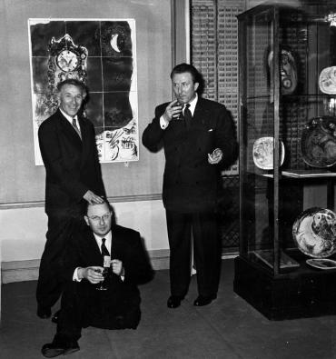 Chagall smiling with hand resting on the head of Clayeux, sitting on the floor, next to Maeght, who is drinking.